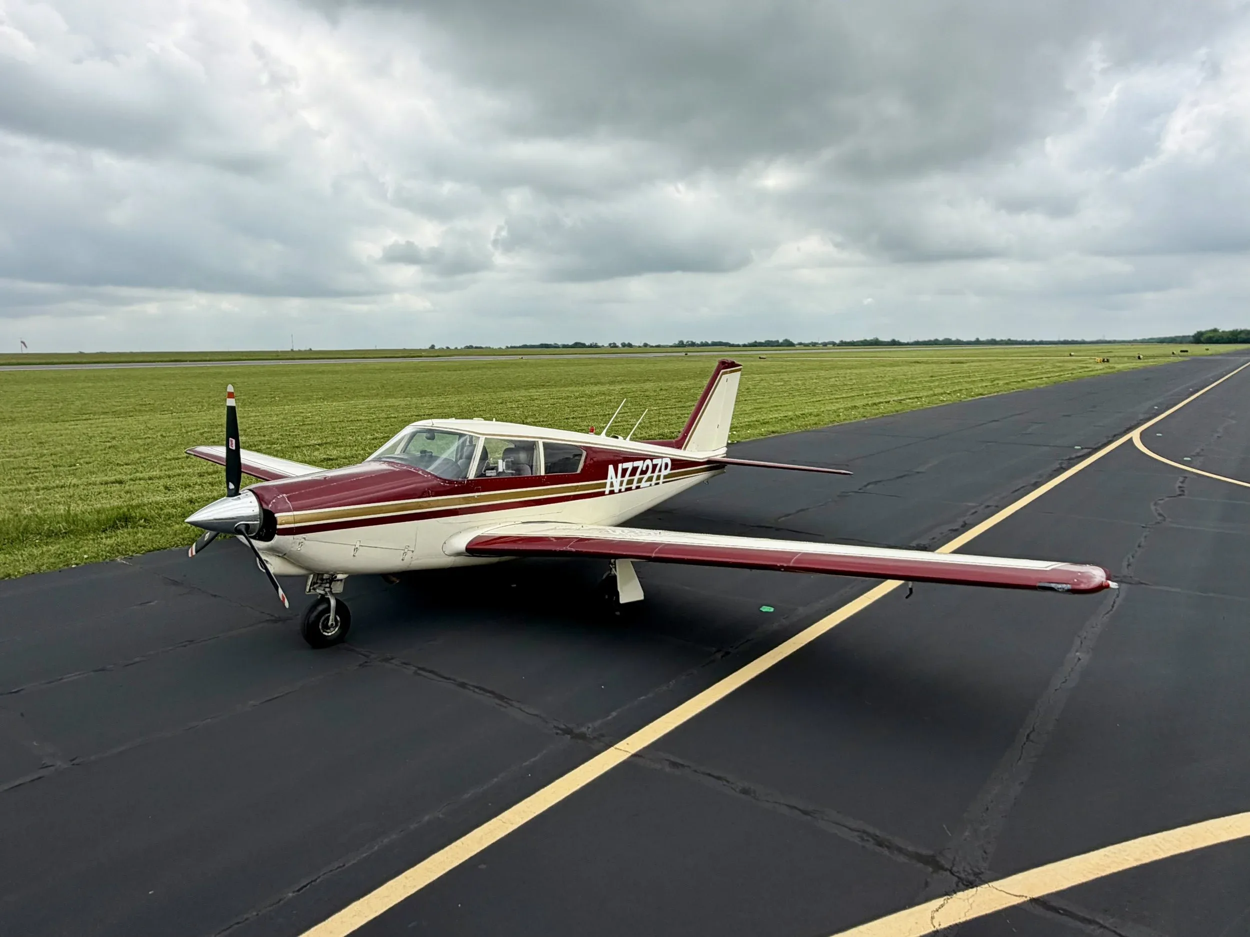 A maroon and white Piper Comanche N7727P parked on an asphalt runway under a cloudy sky.