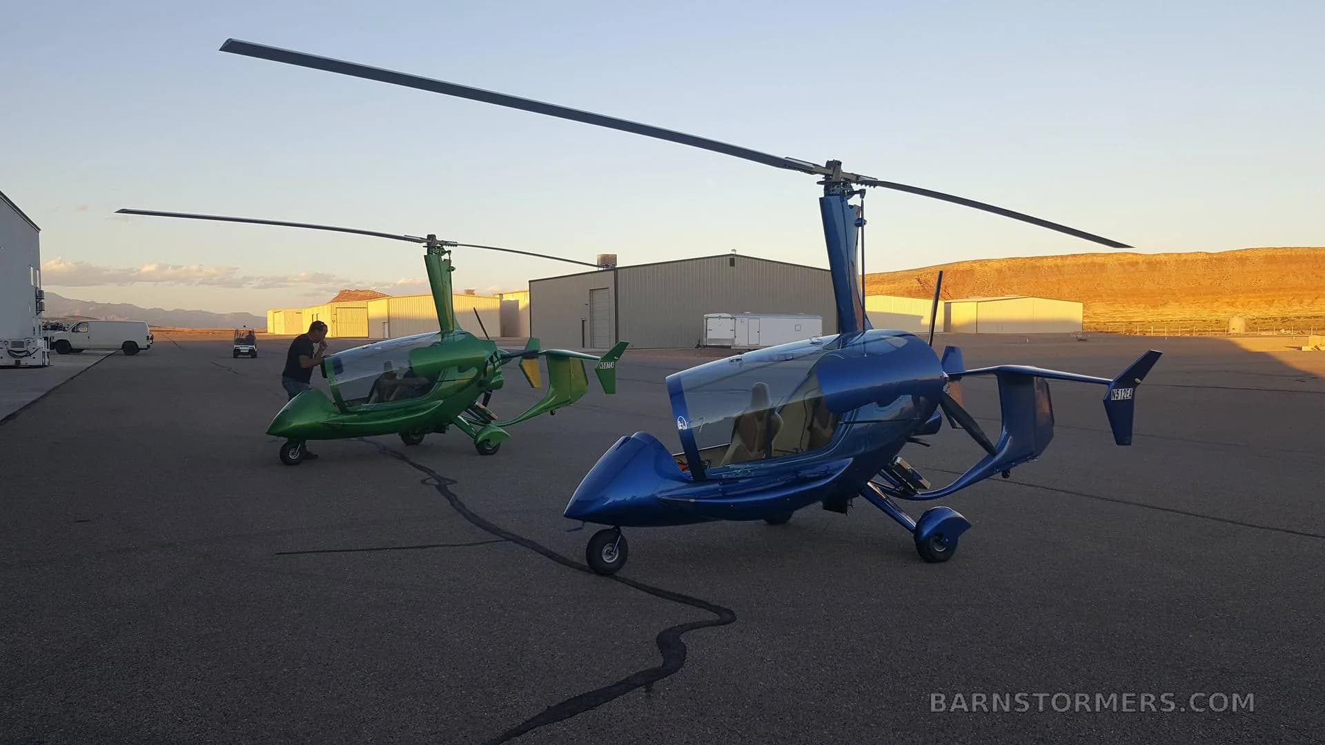 Two enclosed gyrocopters, one blue and one green, are parked on an airfield at sunset with hangars and desert mountains in the background.