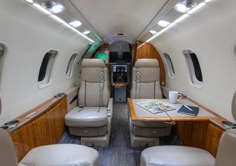 The luxurious cabin interior of a business jet, showing two beige seats facing a table with a magazine and mug.