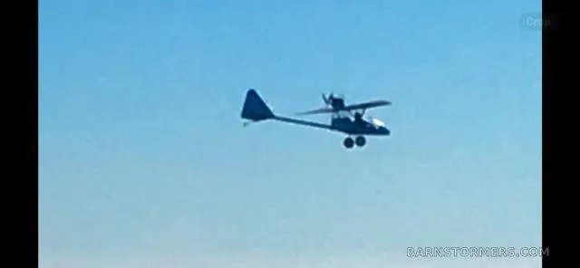 A blue ultralight aircraft with a high wing flying against a clear blue sky.