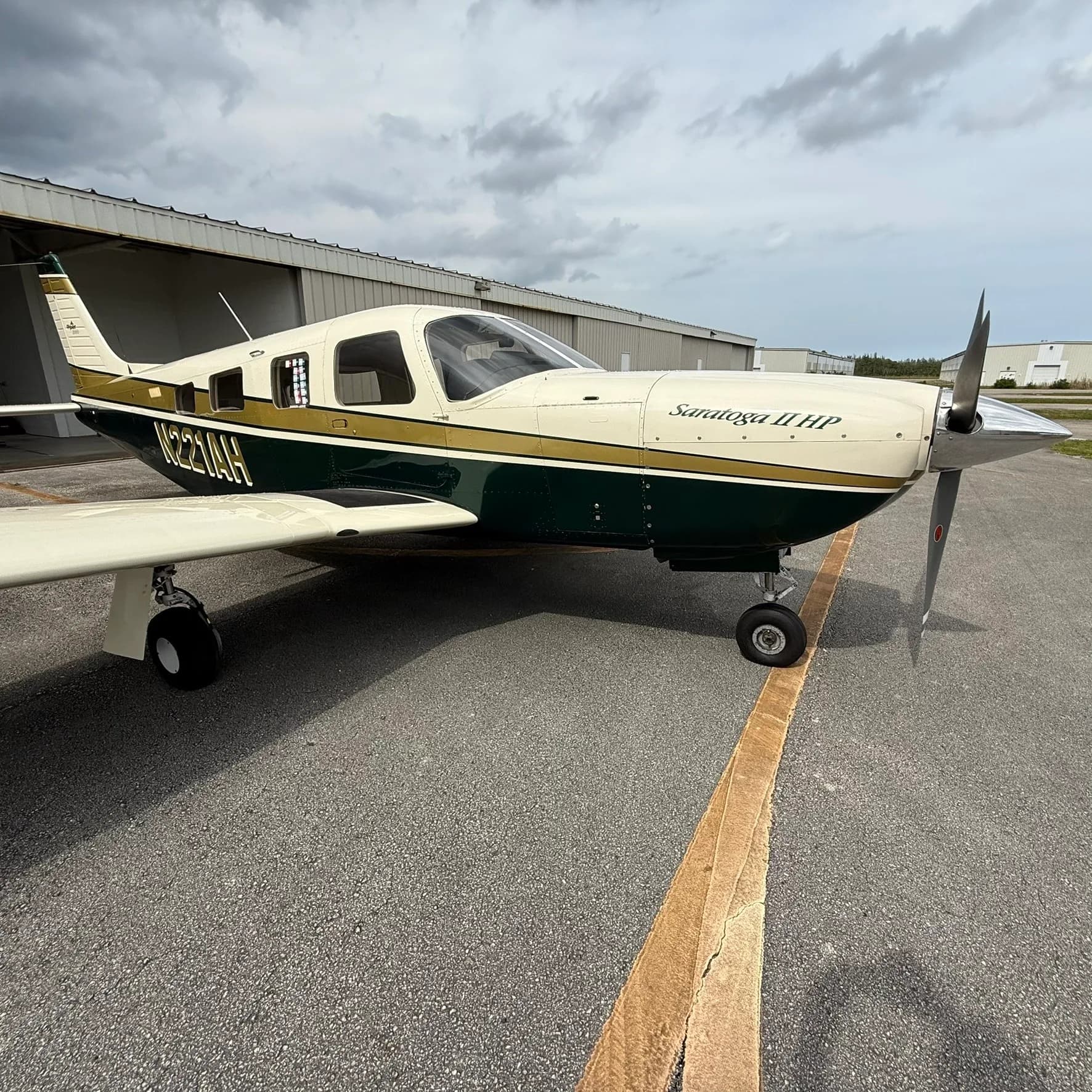 A Piper Saratoga II HP, N221AH, with a white, gold, and dark green paint scheme, parked on an airport tarmac under a cloudy sky.