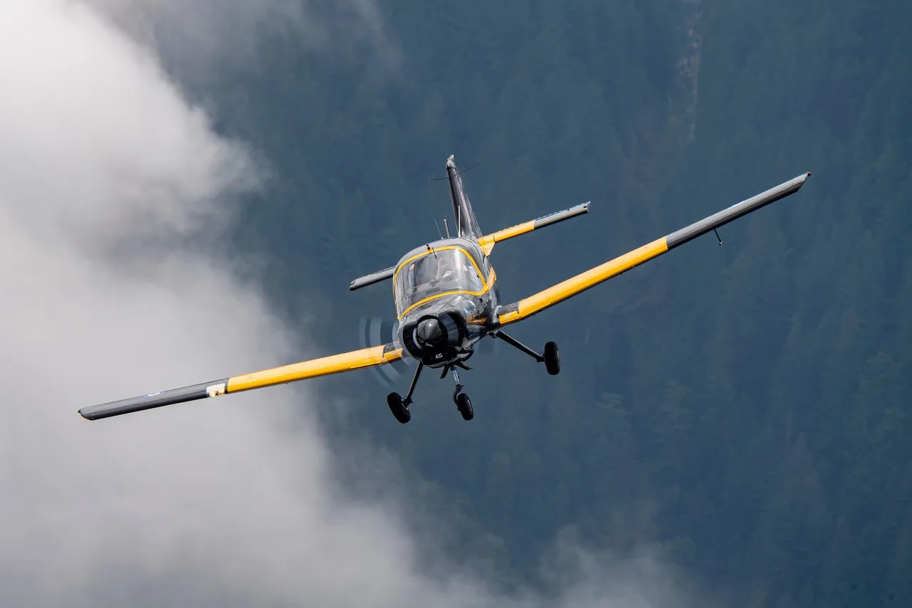 A small black and yellow aircraft with landing gear extended, flying against a cloudy, mountainous background.