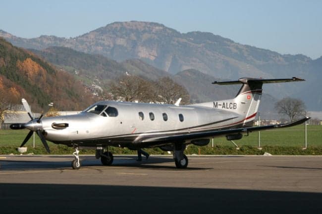 Pilatus PC-12 aircraft with silver paint scheme parked on the tarmac with mountains in the background.