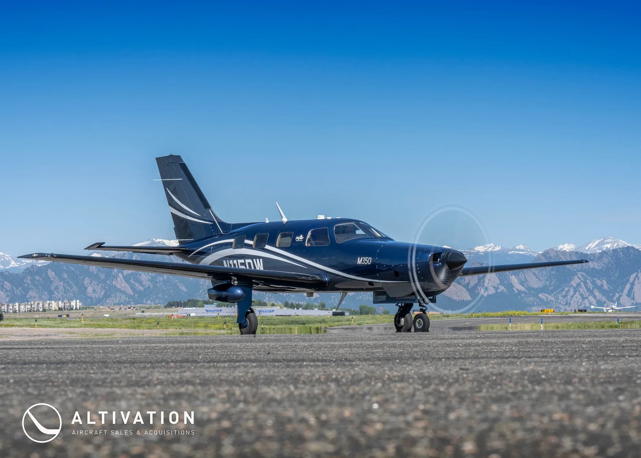 A dark blue Piper M350, tail number N115DW, on a tarmac with mountains and other aircraft in the distance.