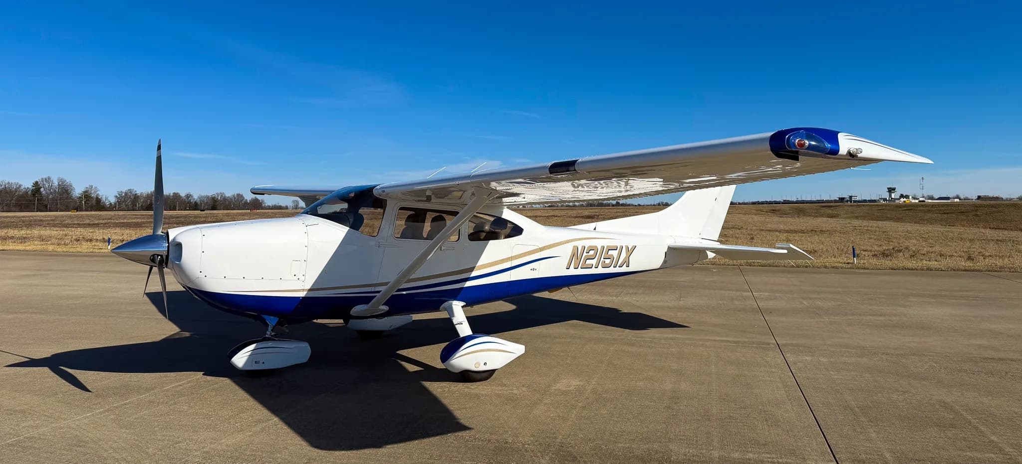 A white and blue Cessna 182 Skylane (N2151X) parked on a tarmac, viewed from the front-right side.