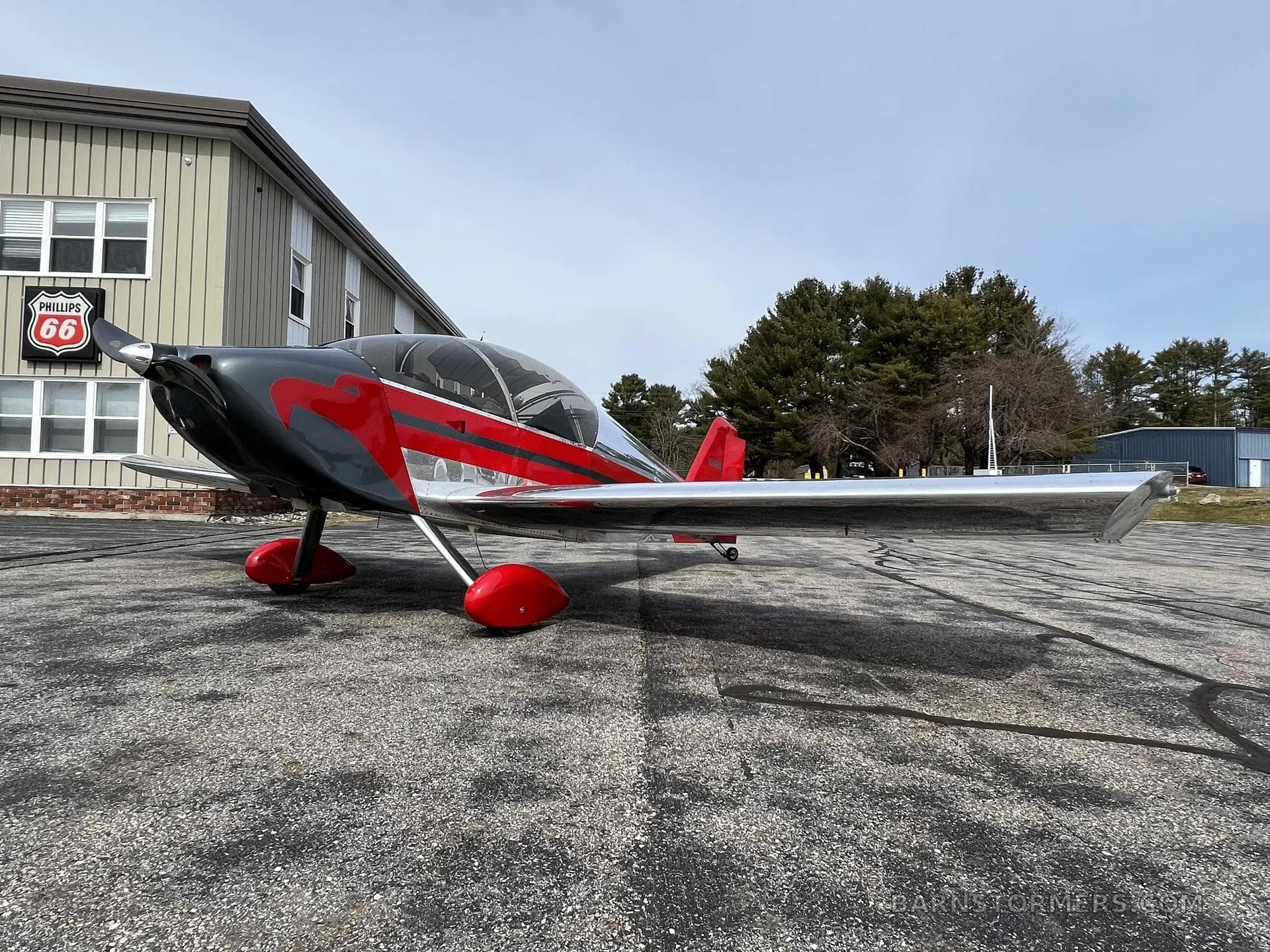 A red and silver experimental aircraft is parked on an asphalt lot next to a building with a Phillips 66 sign.