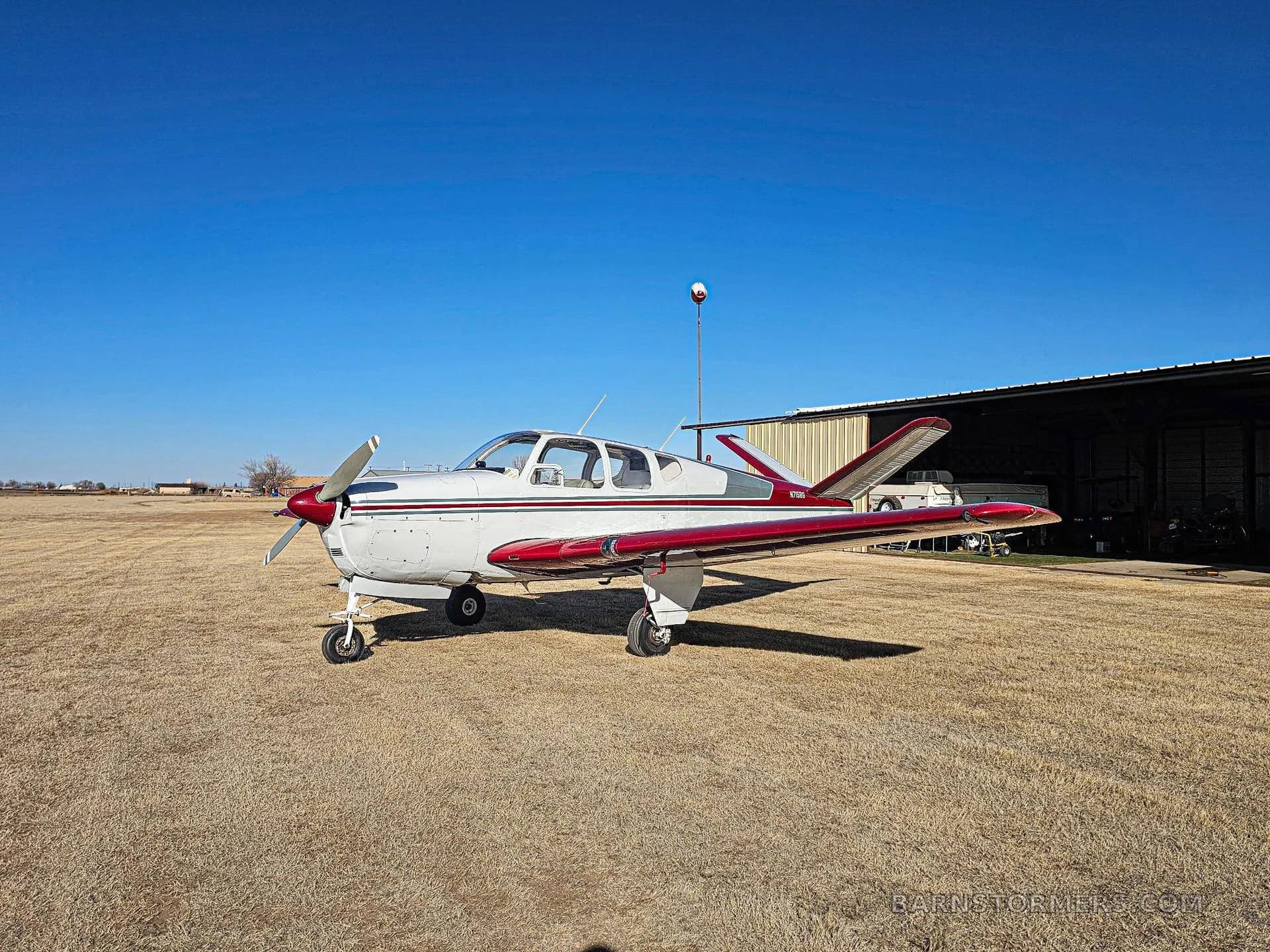 A white and red Mooney M20 series aircraft parked on a grassy field in front of an open hangar under a clear blue sky.