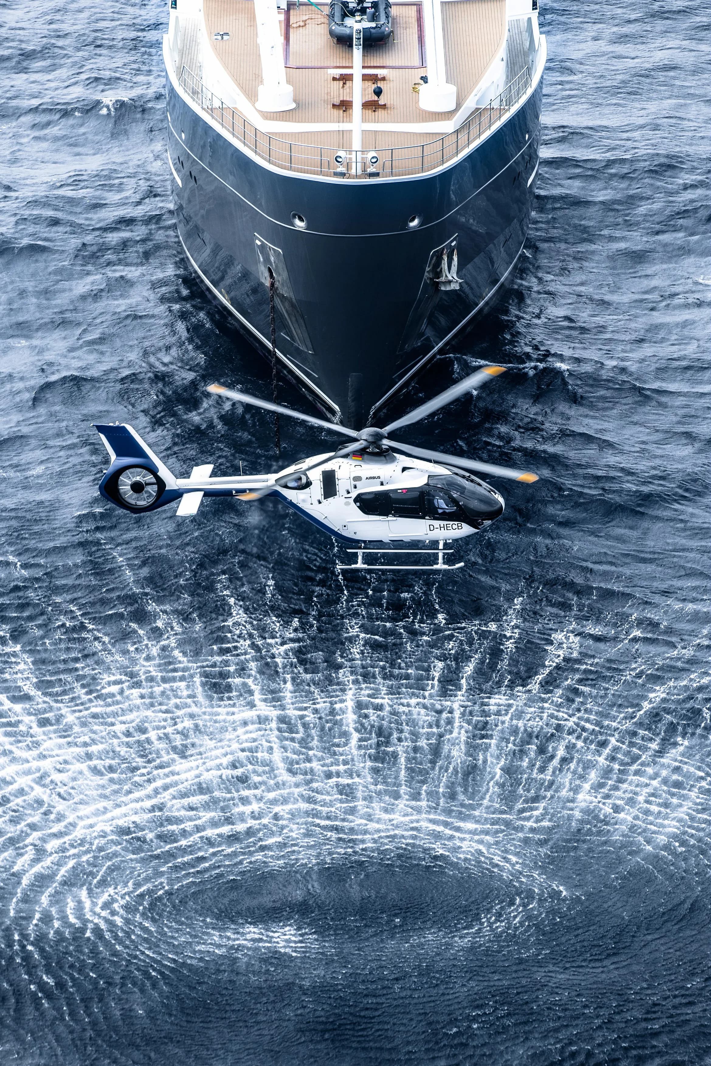 A white and blue Airbus EC135 helicopter flying near a yacht.