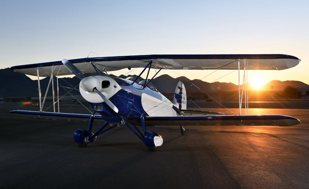 A blue and white biplane, N80JV, is parked on the tarmac with mountains in the background.