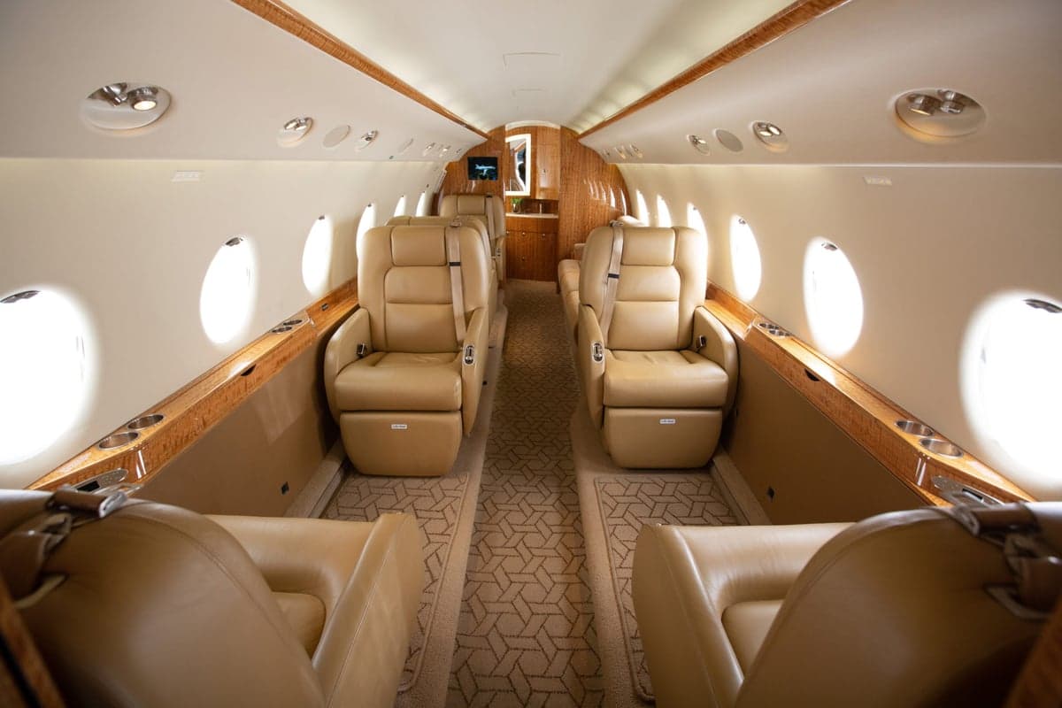 The spacious interior cabin of a private jet with beige leather seats and wood accents.