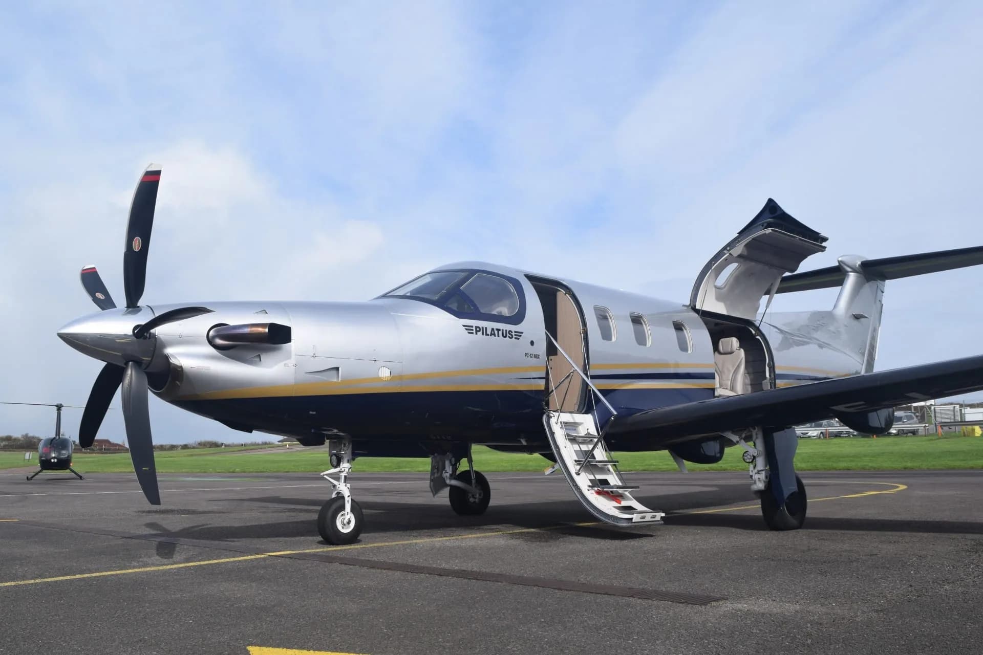 A silver Pilatus PC-12 NGX with blue and gold stripes sits on an airfield tarmac with its doors open, a helicopter visible in the background.