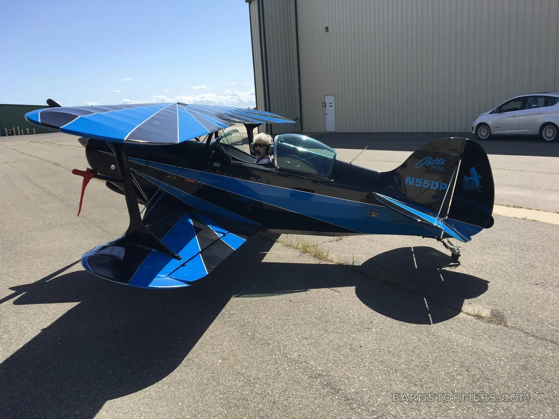A black and blue Pitts Special biplane, tail number N55DP, with a pilot in the cockpit, is parked on an airfield tarmac.