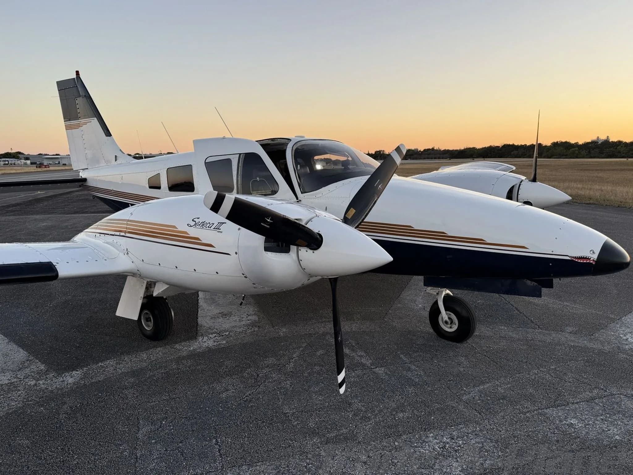 A Piper Seneca III twin-engine aircraft with white and blue paint scheme and gold stripes, parked on an airfield at sunset.