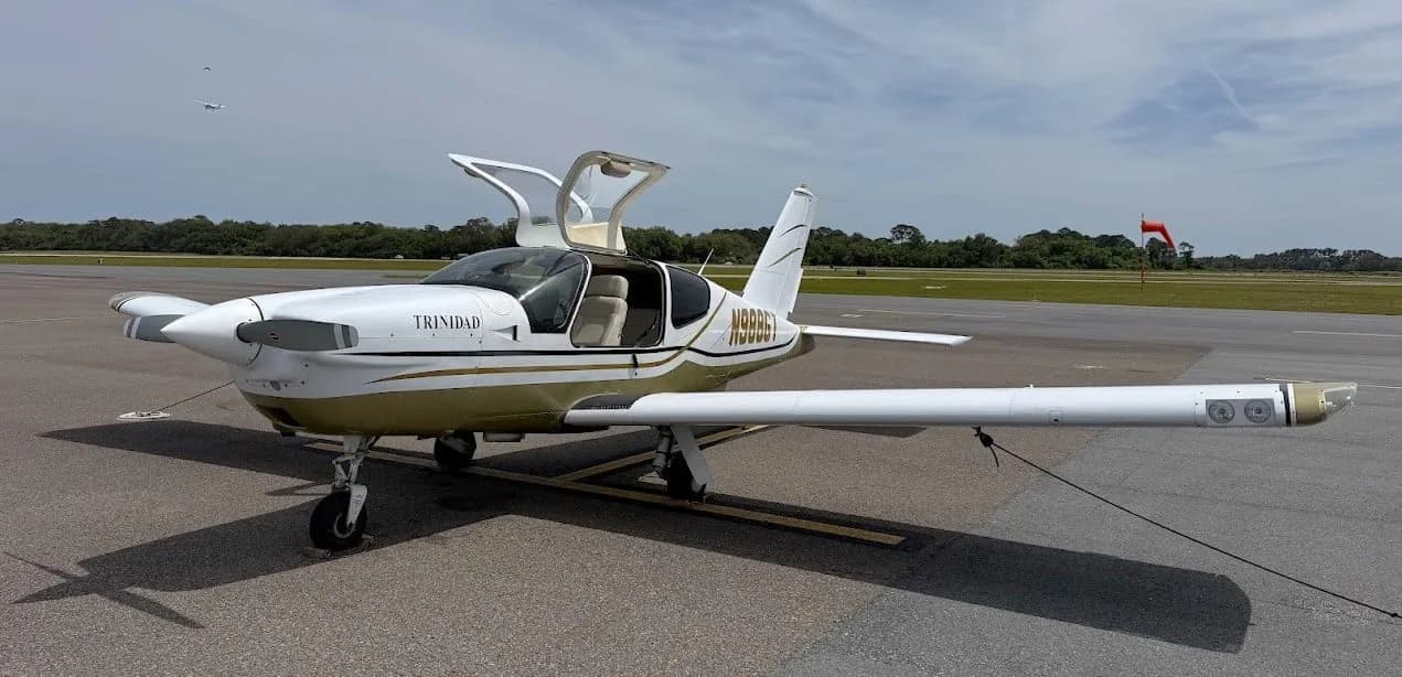 A front-quarter view of the white and gold Socata TB-20 Trinidad aircraft with its doors open on an airfield.