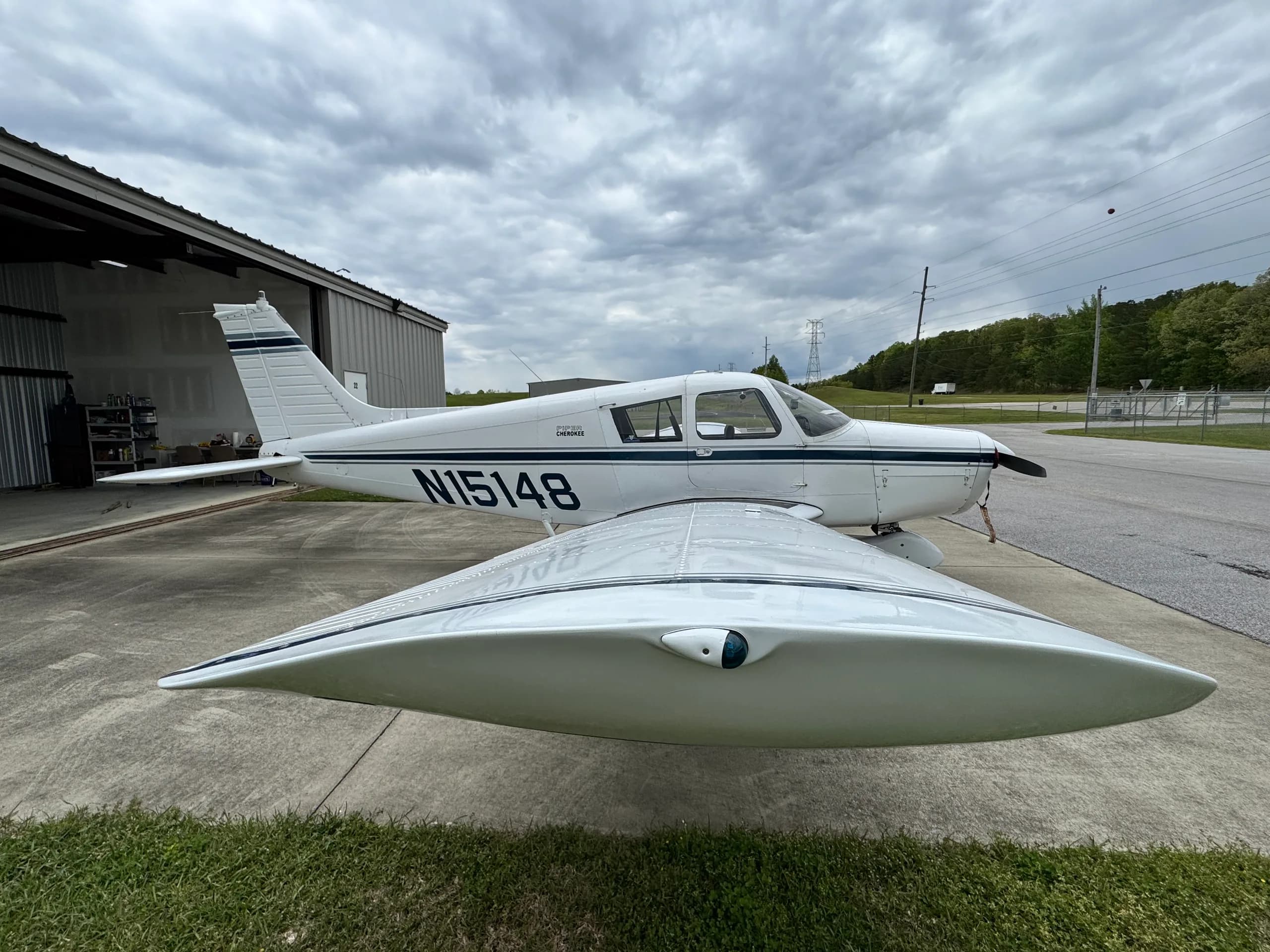 A white single-engine aircraft, N15148, with dark blue stripes, parked on concrete next to a hangar under a cloudy sky.
