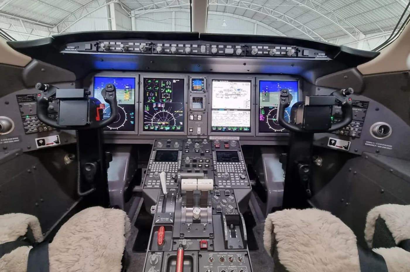 The modern cockpit of a business jet features multiple digital displays, control yokes, and a central control panel, with two sheepskin-covered pilot seats.