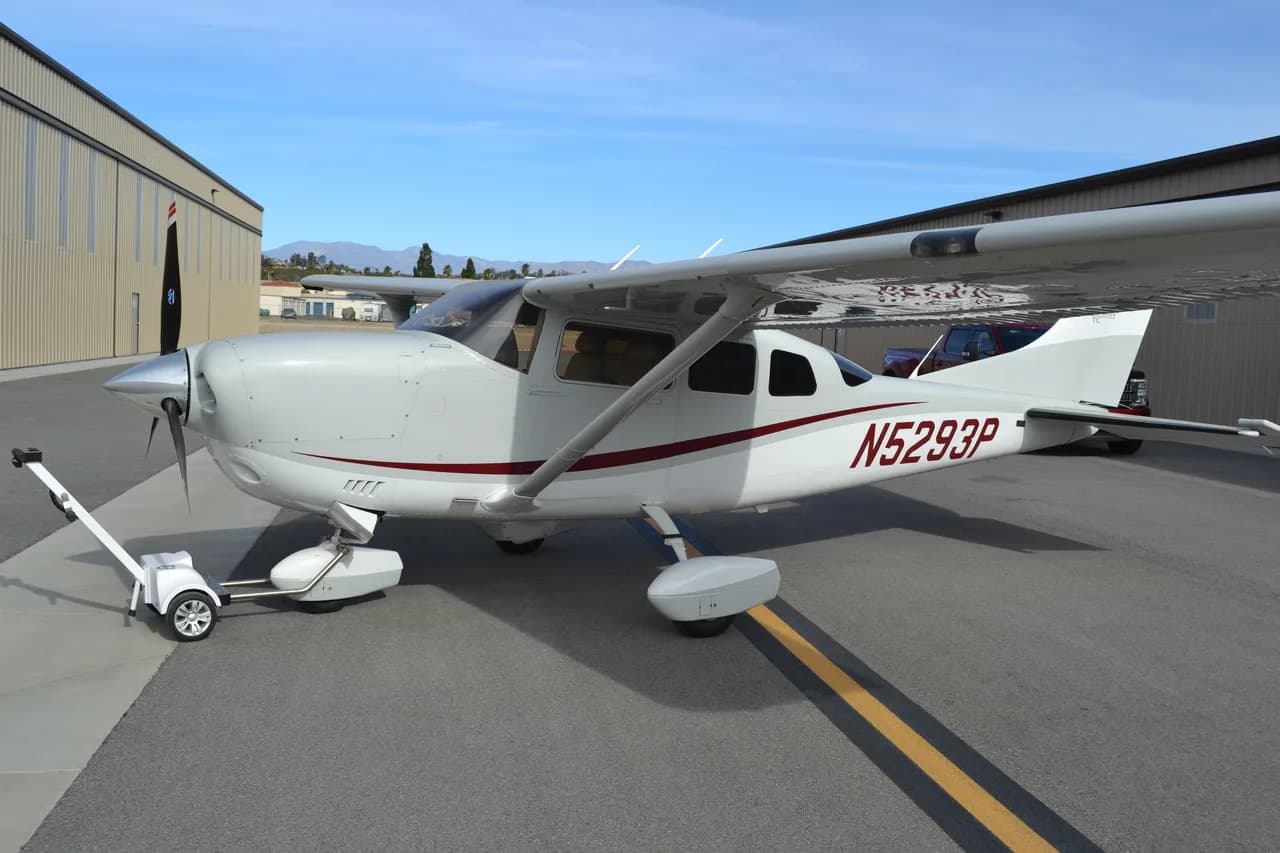 A white single-engine propeller aircraft with red and grey stripes is parked on an asphalt tarmac next to a hangar, with a tow tug attached to its nose wheel.
