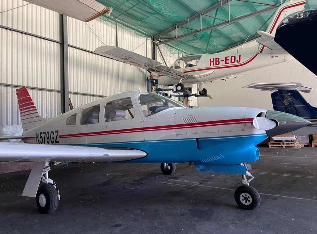 A white and blue Piper PA-28-181 Archer II with red stripes, parked inside a hangar with other aircraft.
