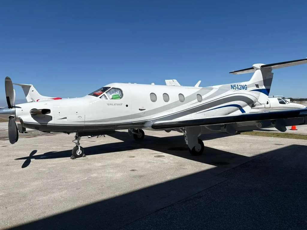A white Pilatus PC-12 NG aircraft with blue and grey stripes, registration N542NG, parked on a tarmac under a clear blue sky.
