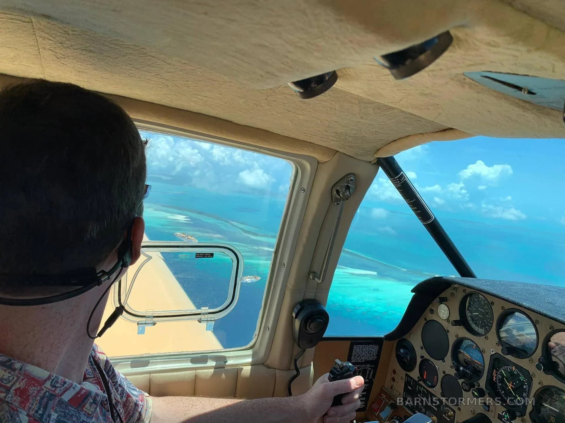 A pilot in the cockpit of a small aircraft, looking out at a stunning view of turquoise ocean and small islands from above.
