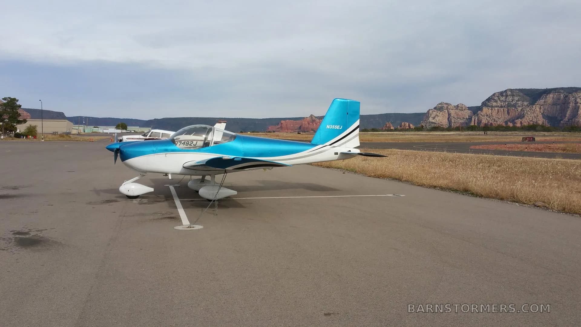 A blue and white single-engine aircraft with registration N355EJ is parked on an airfield, showing the right side with red rock formations in the distance.