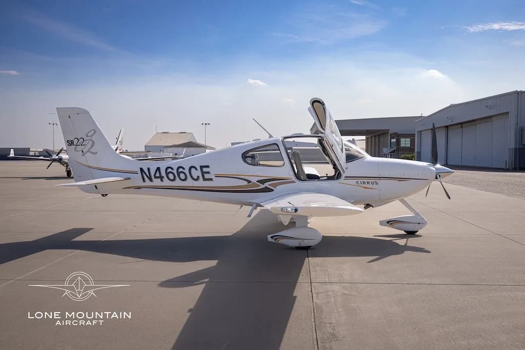A white Cirrus SR22 aircraft with gold and black stripes, registration N466CE, parked on an airfield with its doors open, viewed from the left side.