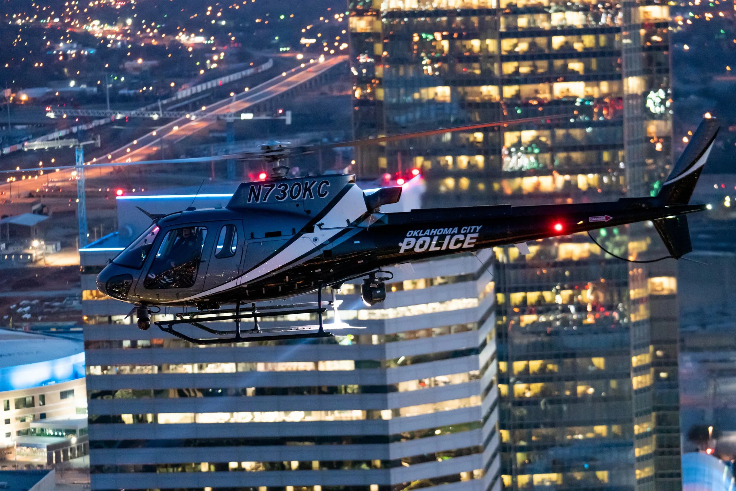 An Oklahoma City Police helicopter flies in front of a city skyline at night.