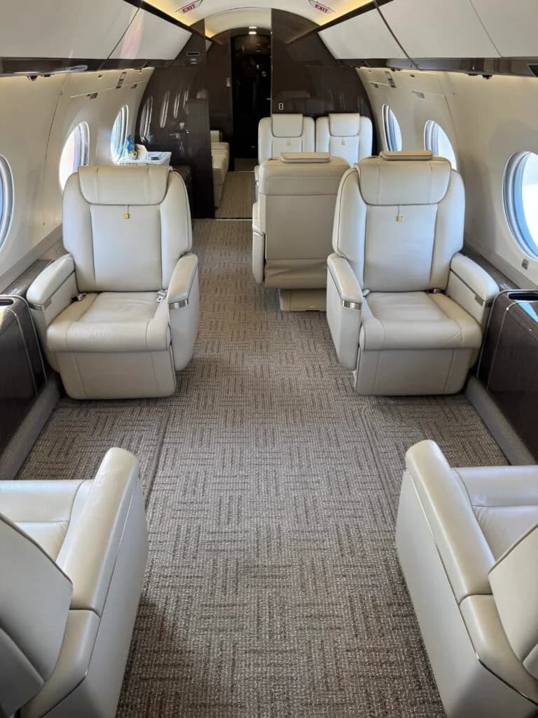 A view down the aisle of a Gulfstream cabin, showing multiple rows of light-colored leather seats and dark wood accents.