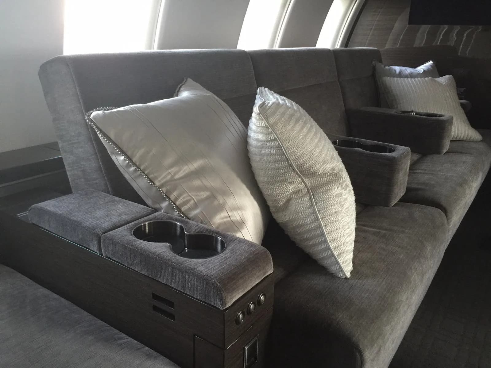 A comfortable grey sofa with white pillows and integrated cup holders inside a private jet cabin.