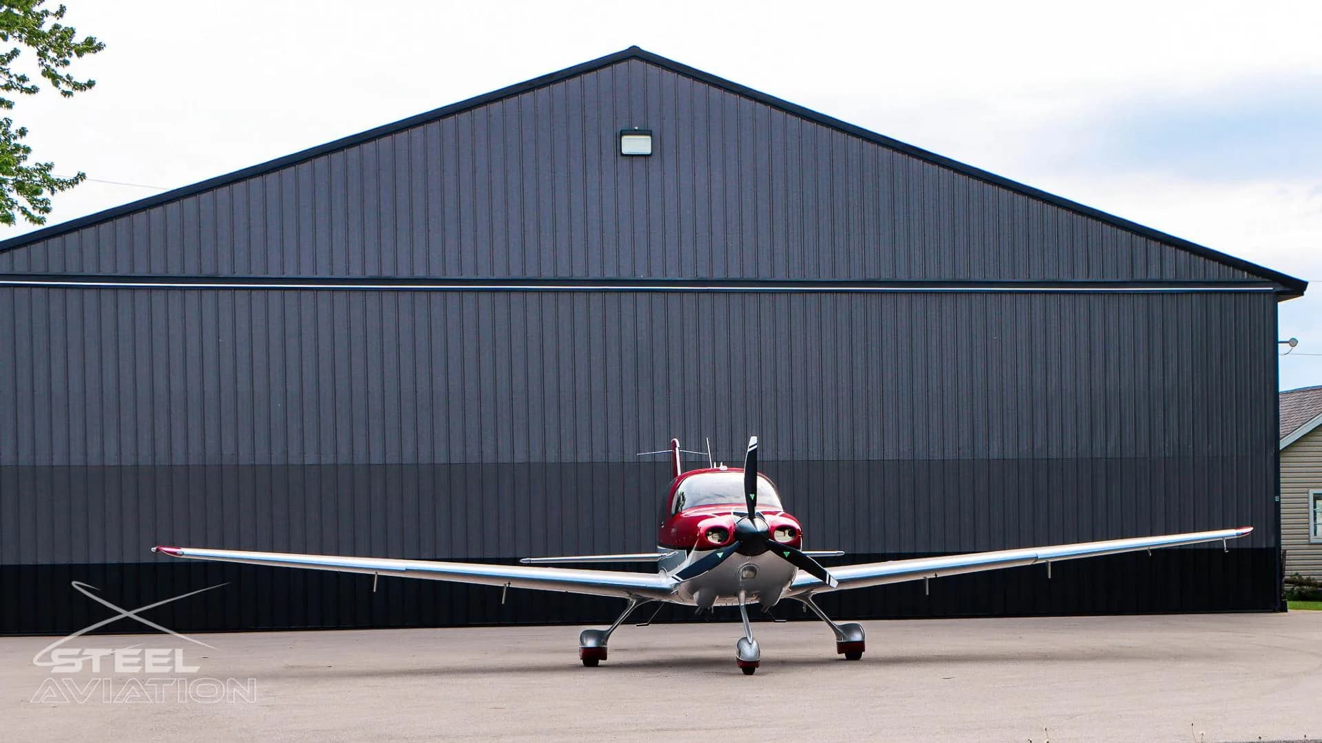A red and white Cirrus SR22 aircraft is parked head-on in front of a dark gray hangar under a cloudy sky.