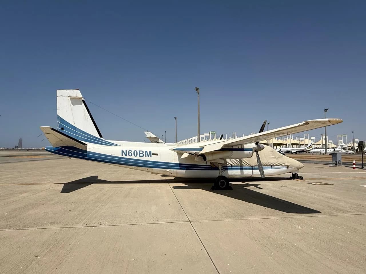 A white and blue twin-engine turboprop aircraft, registration N60BM, is parked on an airfield under a clear sky, with its nose covered.