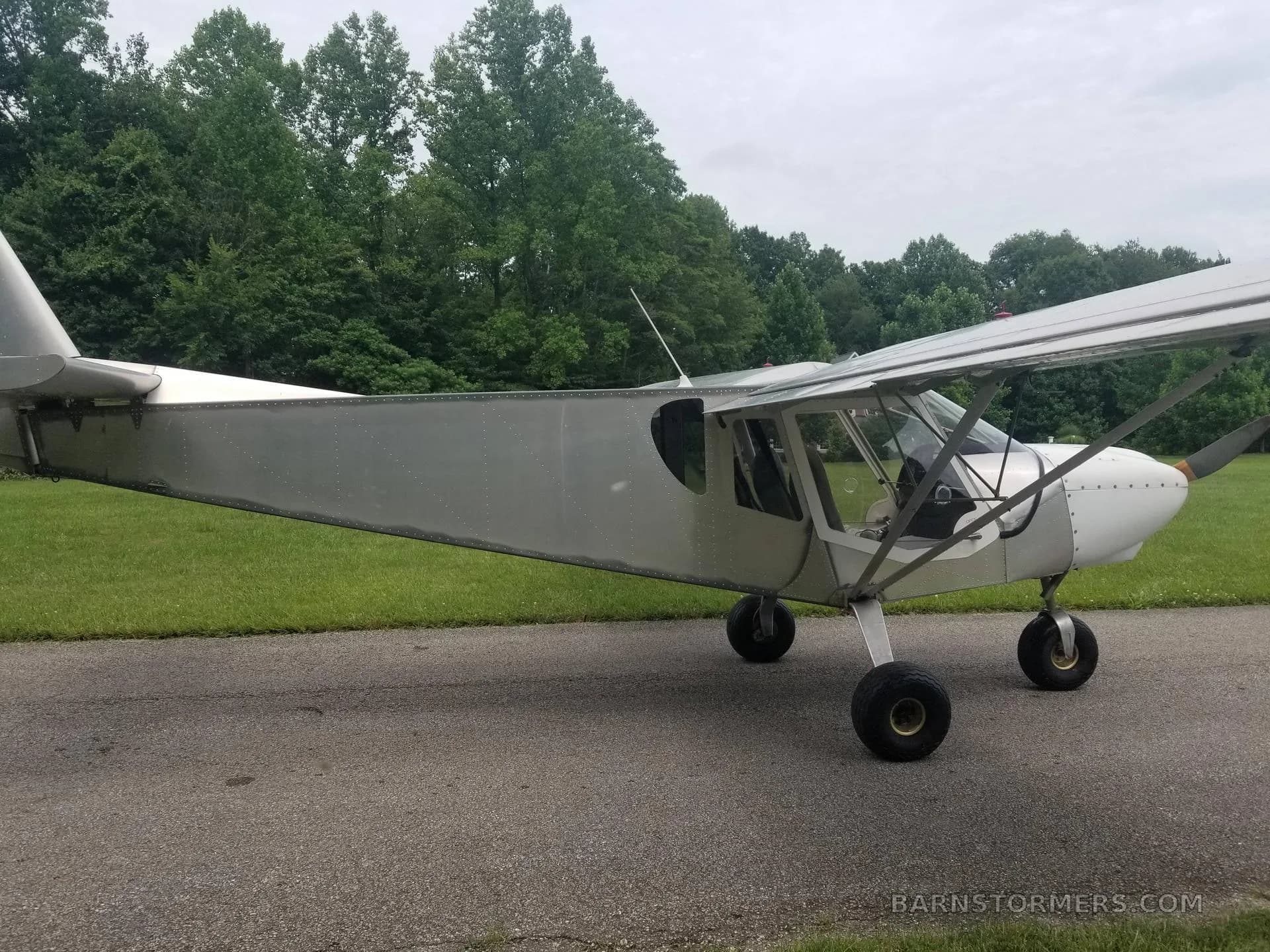 A side view of a light gray ultralight aircraft with a high wing, parked on a paved path next to green grass and trees.