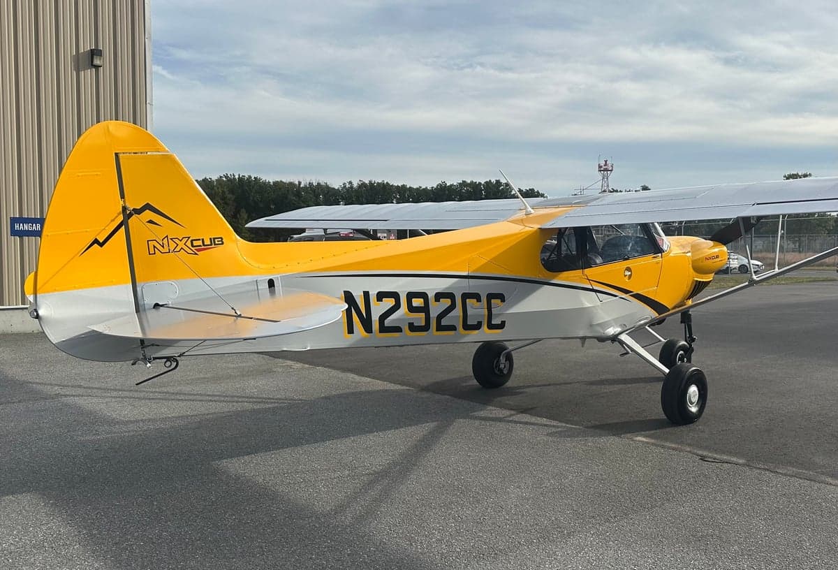 A yellow and gray NX Cub aircraft, N292CC, parked on an asphalt tarmac with a hangar in the background.