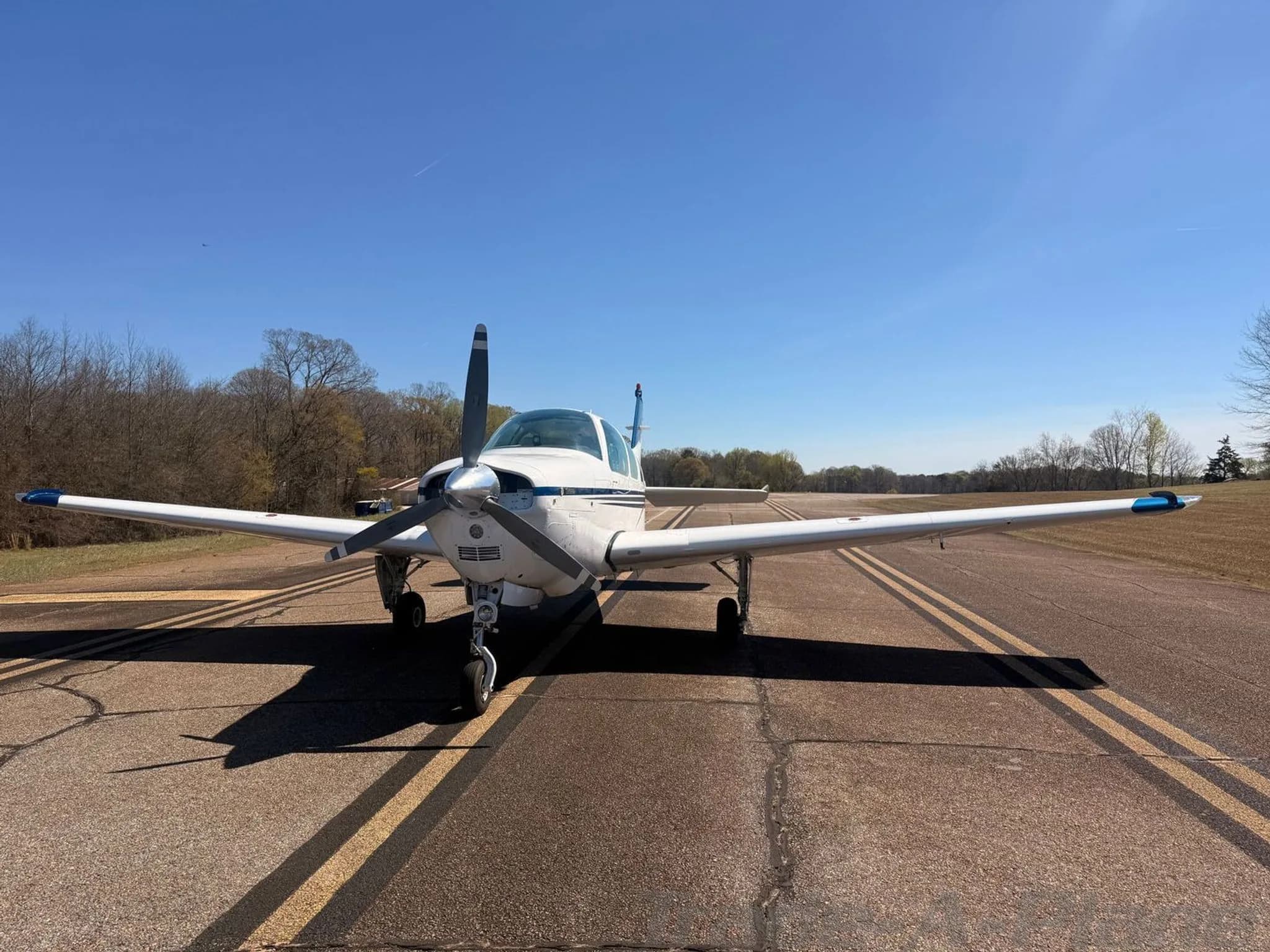 A front view of the white Beechcraft Bonanza N222CR with blue and gold stripes parked on an asphalt runway.