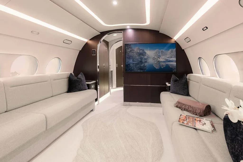 A private jet cabin featuring two long, light grey sofas facing each other, with a large TV screen displaying a winter scene.