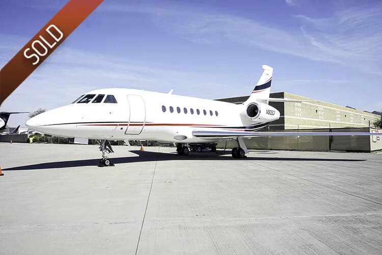 A white Dassault Falcon business jet with red and blue stripes is parked on an airfield under a clear sky.