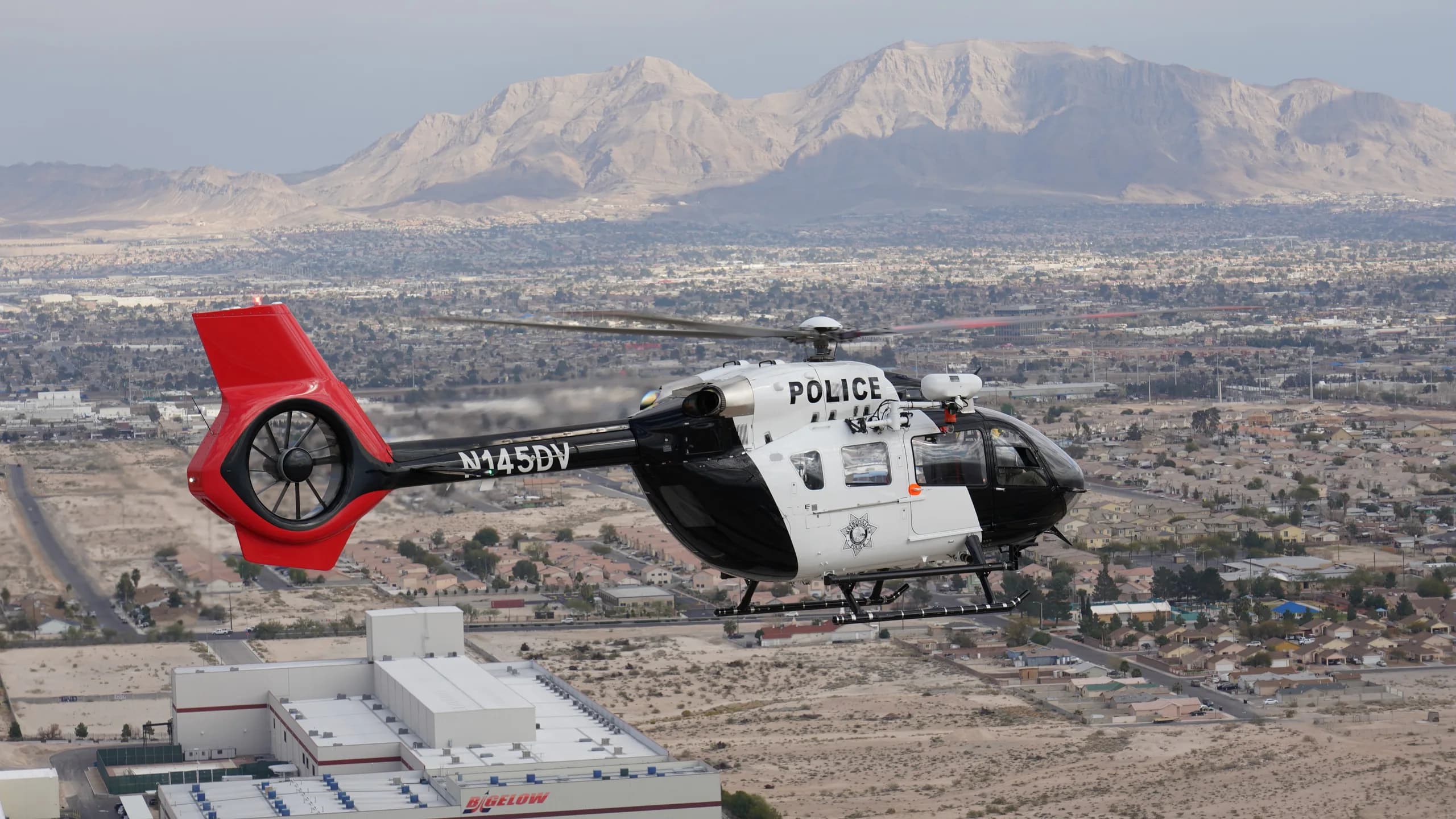 A black and white police helicopter flying over a city with mountains in the background.