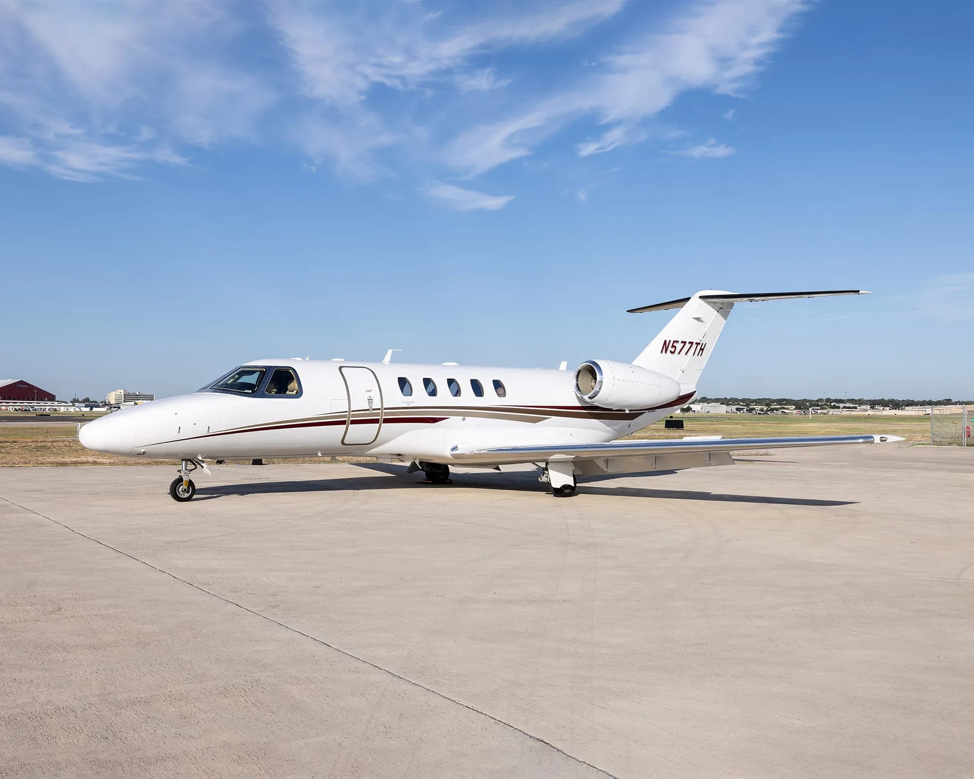 A white business jet with brown and gold accent stripes is parked on an airport ramp under a blue sky.