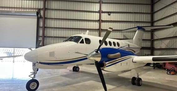 A white twin-turboprop aircraft with blue and gold stripes on the fuselage and engine nacelles, parked inside a hangar, viewed from the front-left.