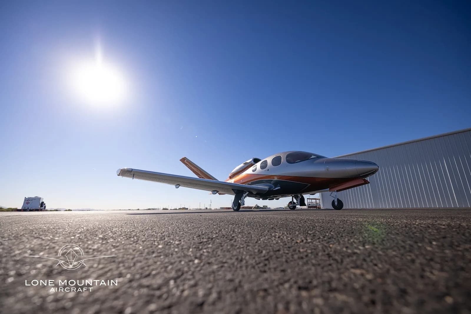 A low-angle view of a silver and orange Cirrus Vision Jet (SF50) parked on an asphalt tarmac under a bright sky.