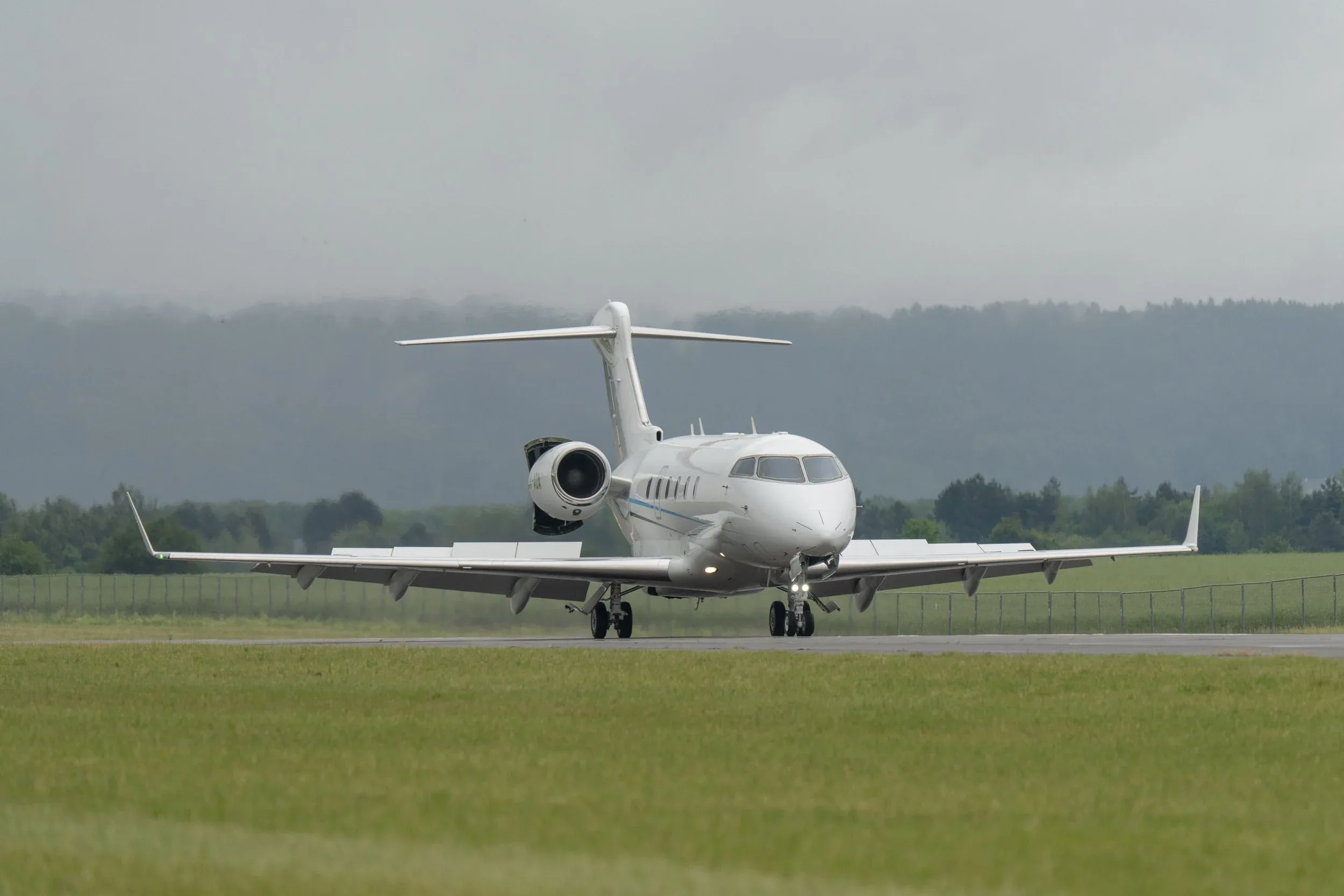 A white business jet with a blue stripe is on a runway, facing forward, with green grass and misty hills in the background.