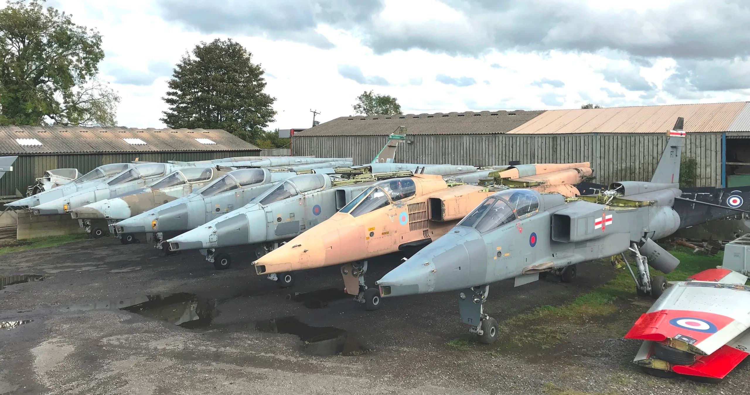 A line of several military jet aircraft, possibly Jaguars, in various paint schemes and conditions, parked outdoors near hangars.