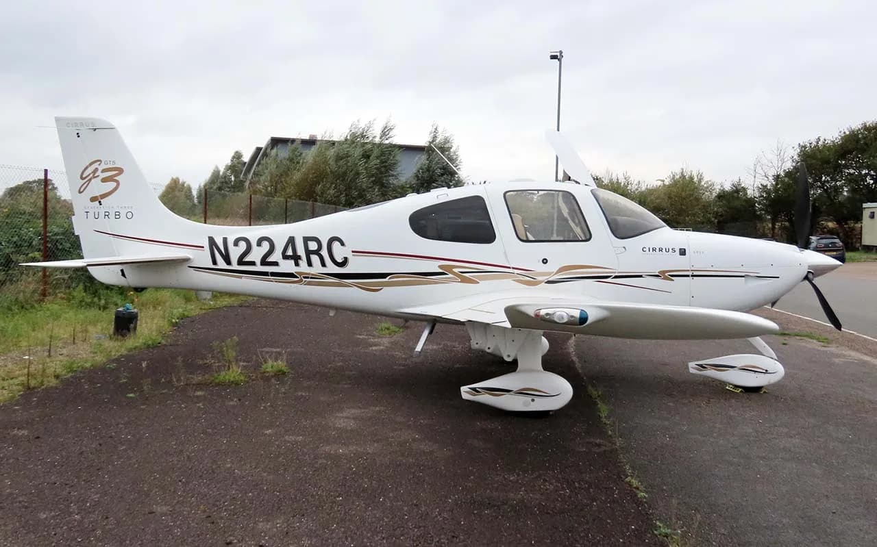 A white Cirrus SR22T (N224RC) with brown and black stripes parked on an asphalt surface next to a grassy area.