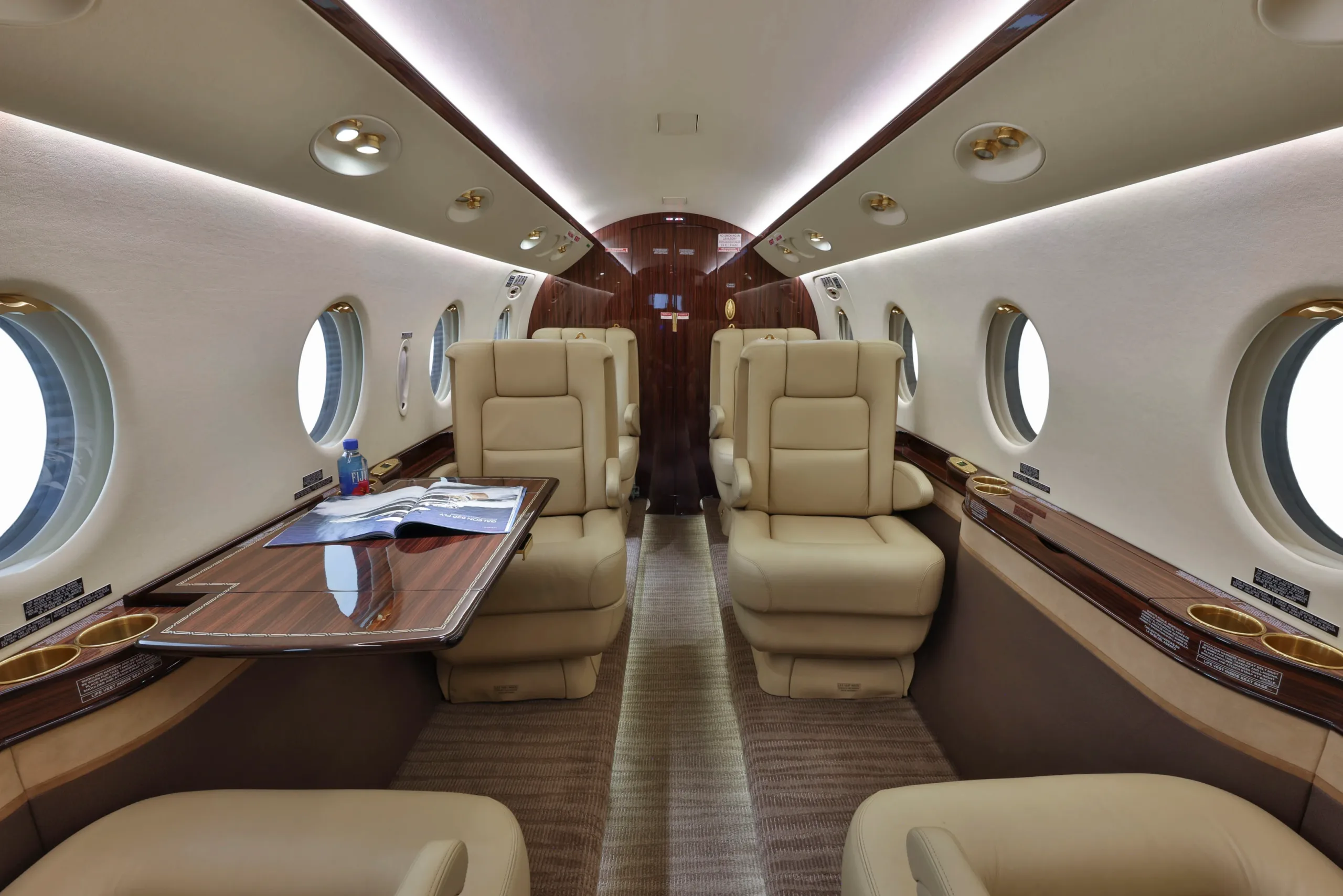A wide view of a private jet cabin interior with beige leather seating, a deployed wooden table, and large windows.