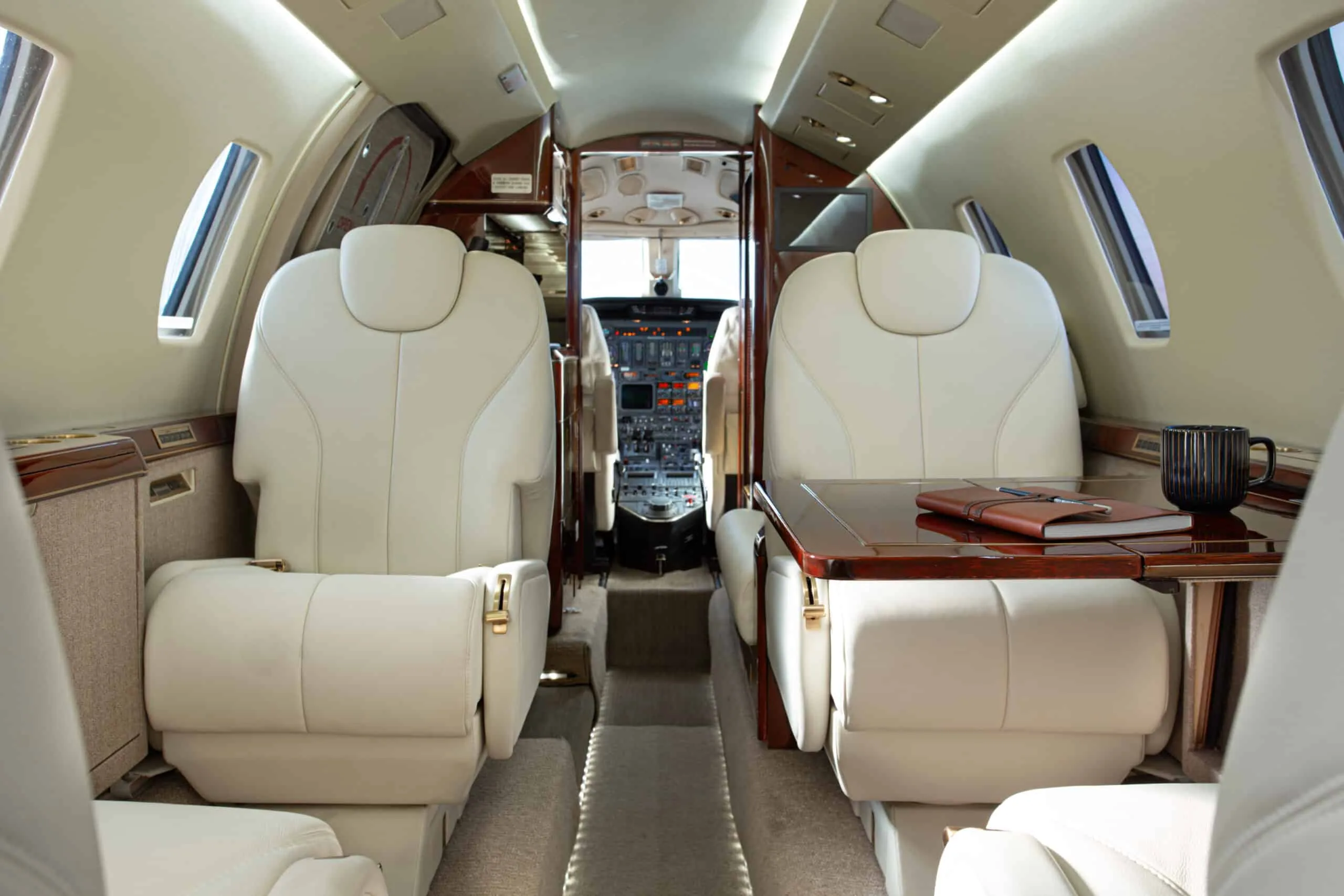 View from the rear of a luxurious aircraft cabin with cream leather seats, a polished wooden table, and a glimpse of the cockpit.