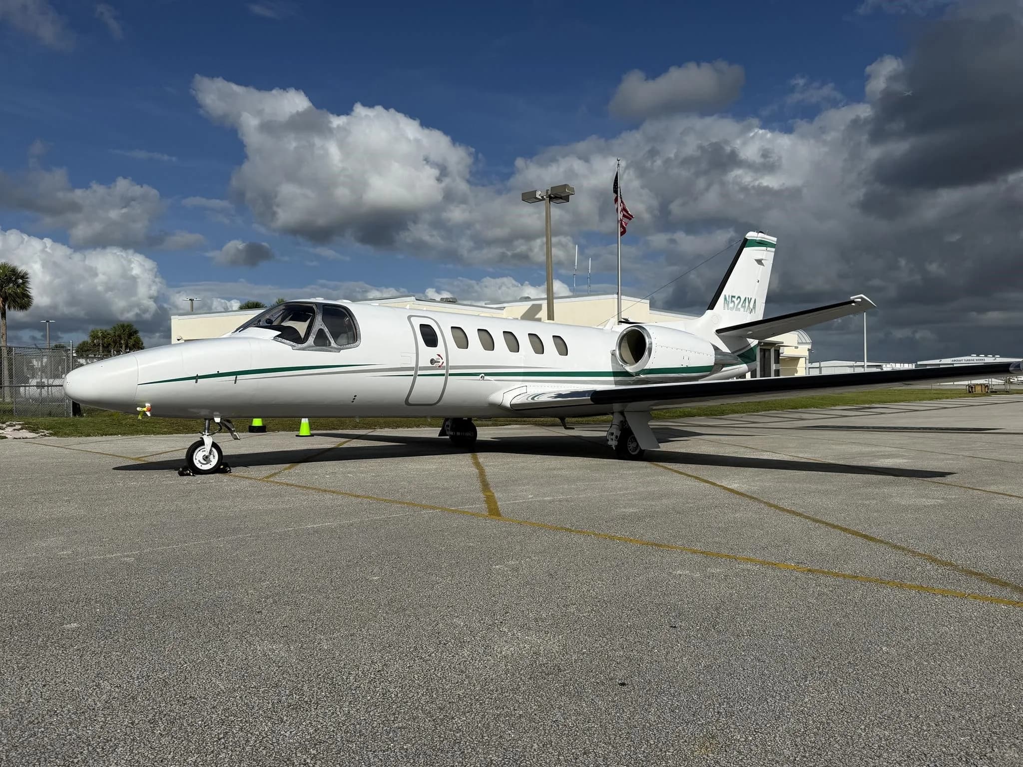 A white Cessna CitationJet with green and grey stripes is parked on an asphalt tarmac under a partly cloudy sky.