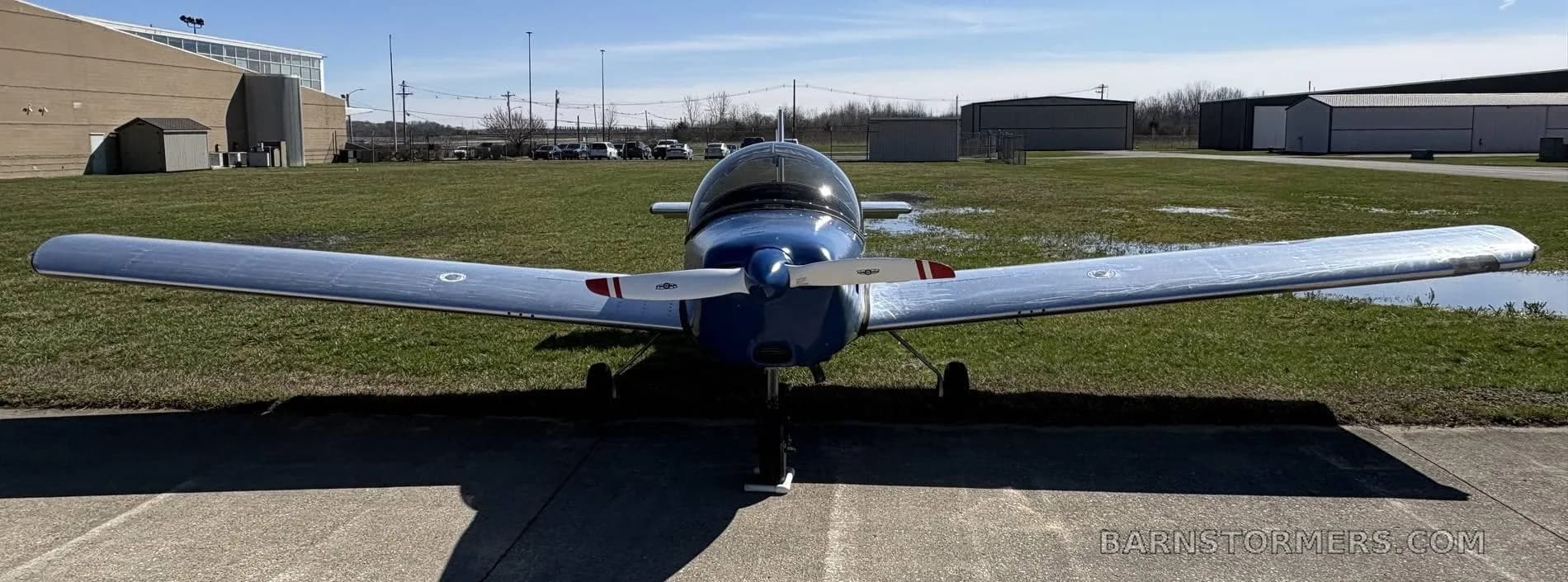 A blue and polished aluminum single-engine aircraft with a propeller, viewed head-on from the front on an airfield.