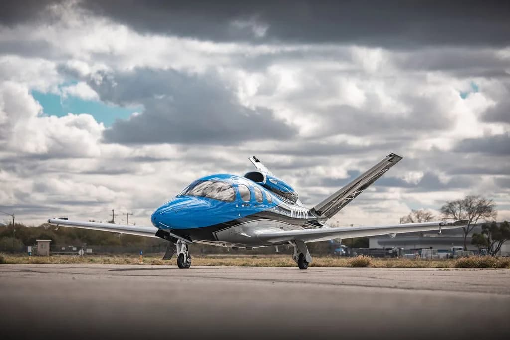 A blue and silver Cirrus Vision Jet parked on an airfield under a cloudy sky.