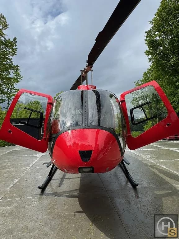 Front view of a red helicopter with its doors open, parked on a wet concrete surface outdoors.