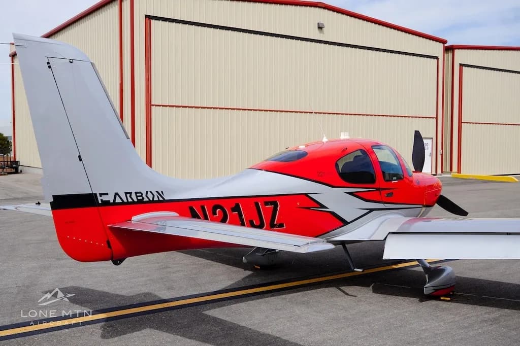 A rear-side view of a red and silver Cirrus SR22 aircraft parked on an asphalt ramp next to a hangar.